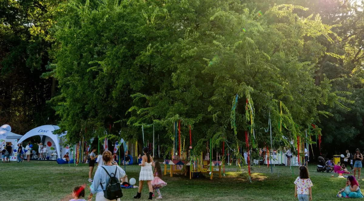 In imagine un copac mare, verde, in mijlocul unei pajiști cu multi oameni, de crengile lui sunt agăgate mai multe panglici colorate