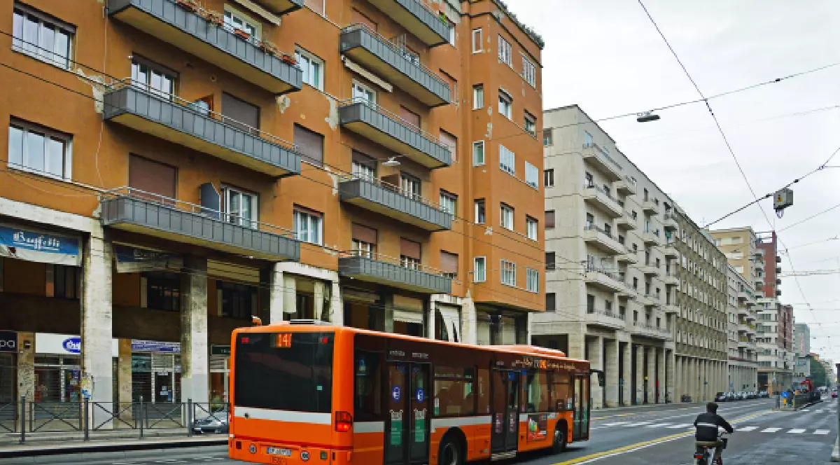 Imaginea arată o stradă dintr-un oraș, cu clădiri cu mai multe etaje pe ambele părți. Un autobuz portocaliu se deplasează pe banda din dreapta, iar pe spatele său este vizibil numărul 14. Clădirile din stânga au o culoare cărămizie și balcoane gri. Pe dreapta, clădirile sunt mai deschise la culoare. Cerul este înnorat, iar pe stradă se văd un biciclist și linii aeriene, sugerând existența unui sistem de transport public electric.