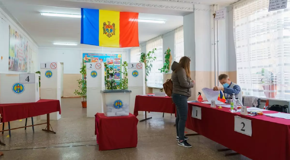 O femeie se află într-o secție de vot în Republica Moldova și așteaptă să fie găsită pe listele electorale de un funcționar, pentru a verifica prezența ei pe lista alegătorilor, înainte de a-i înmâna buletinul de vot