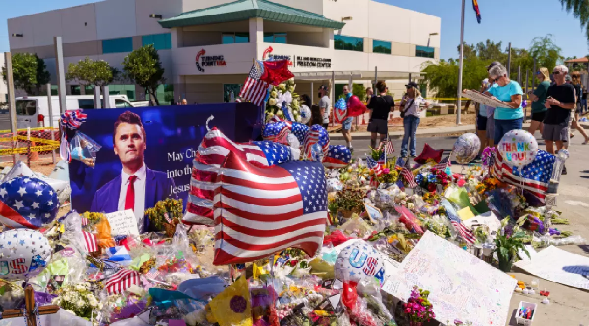 Imaginea prezintă un memorial improvizat în fața clădirii Turning Point USA. În centrul atenției se află o grămadă mare de obiecte, majoritatea având tematica steagului american: baloane în formă de stea, inimă și de steag, precum și mici drapele americane înfipte printre flori, lumânări și diverse mesaje scrise. Un balon specific are scris mesajul 