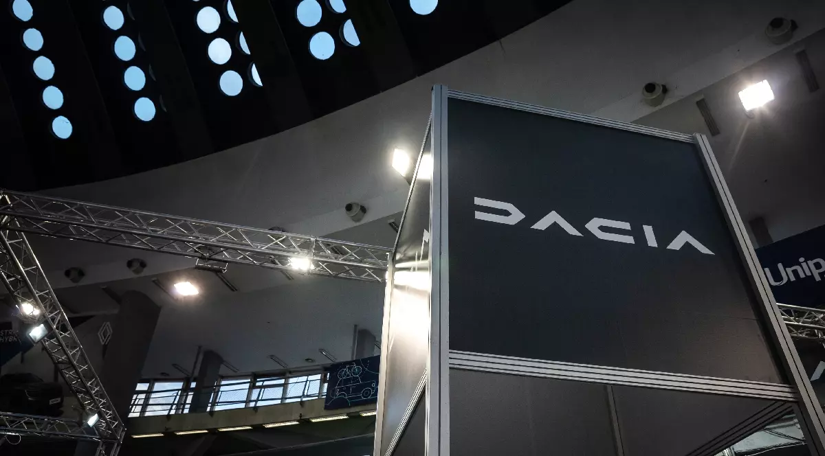 Imaginea surprinde un stand al mărcii de mașini Dacia, probabil la un târg sau un salon auto, judecând după structura metalică a standului și luminile de spot. Pe un panou mare, de culoare închisă, este scris cuvântul "DACIA" cu litere albe, stilizate. Deasupra standului se observă o structură arhitecturală a tavanului, compusă dintr-un rând de ferestre circulare, iluminate, ce lasă să pătrundă o lumină difuză, dar și structuri metalice de susținere și reflectoare, specifice unui spațiu expozițional.