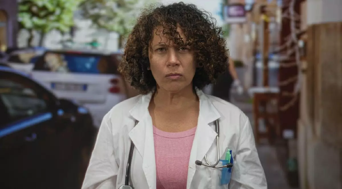 Femeie stând pe o stradă îngustă dintr-un oraș, îmbrăcată într-un halat alb, cu stetoscopul la gât, cu o expresie serioasă, privind în față.