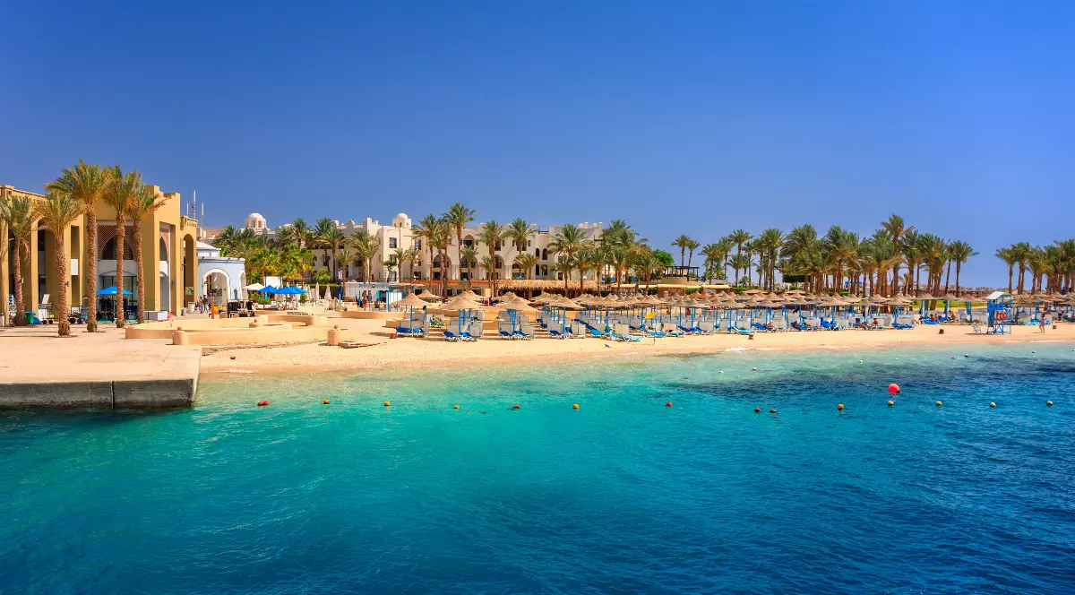 O plajă dintr-un resort din Egipt, cu nisip alb, mărginita de palmieri și de o clădire mare, în stil mediteranean. Marea, de un albastru intens, are ape cristaline în apropierea țărmului, în timp ce în larg se observă o nuanță mai închisă, de un albastru marin profund.