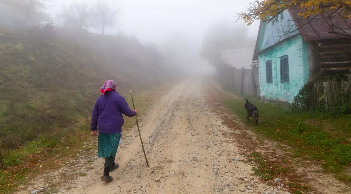 Femeie bătrână, îmbrăcată cu o fustă verde și un pulover albastru, cu un batic roșu pe cap, pe uliță cu un băț în mână, lângă o casă vopsită verde