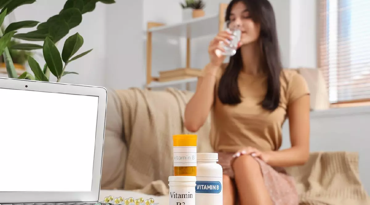 Tânără luând pastile acasă, pe masă se văd recipiente cu diferite vitamine B și laptopul