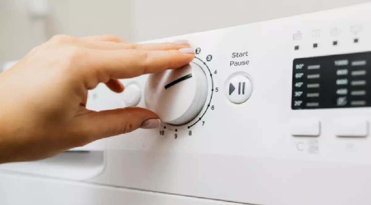 Imaginea surprinde un prim-plan al panoului de control al unei mașini de spălat moderne de culoare albă, unde o mână cu manichiură delicată rotește un buton selector mare pentru a alege un program. Sunt vizibile și alte butoane, inclusiv cel de Start/Pauză, precum și un ecran digital ce afișează opțiuni de temperatură și clătire.