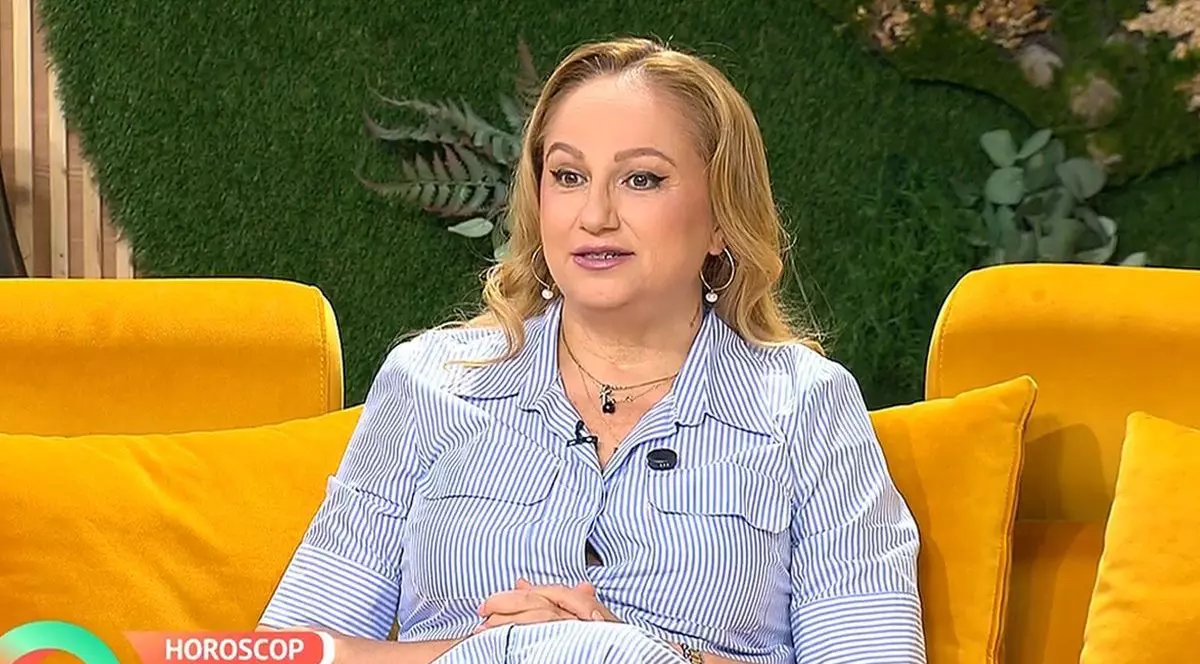 Astrologul Cristina Demetrescu, îmbrăcată cu o rochie cu dungi alb-albastru, fotografiată surprinsă, în timp ce stă pe o canapea portocalie, în studioul unei emisiuni tv, București, 15 septembrie 2025