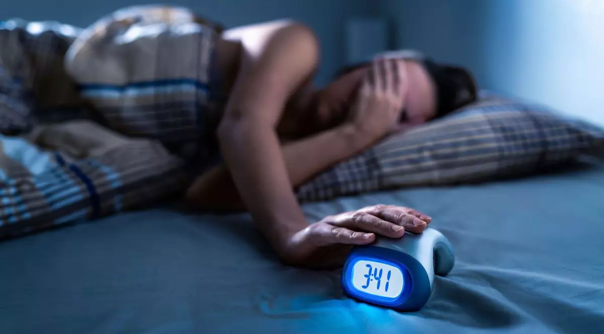 O femeie care este culcată în pat doarme și își acoperă fața cu o mână, iar cealaltă mână este pusă pe ceasul care este în pat lângă ea și arată ora 3:41. Are capul pe pernă și este învelită cu o pătură