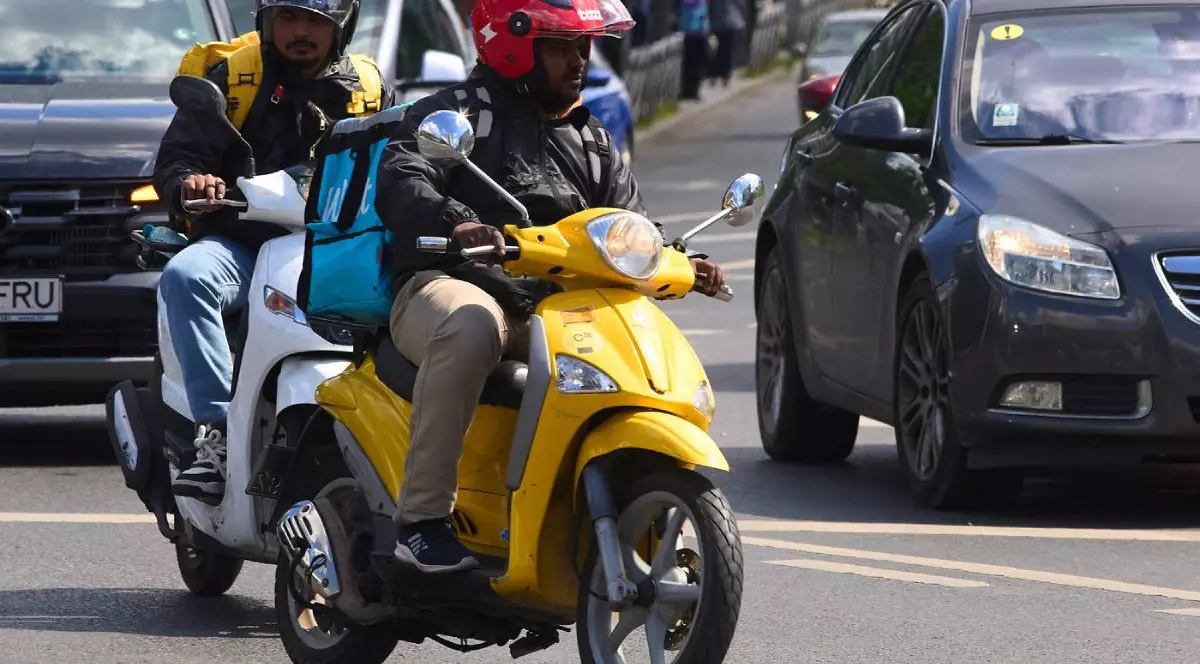 Doi livratori de mâncare, unul pe un scuter galben, și altul pe un scuter alb-albastru, în trafic, pe o stradă din București