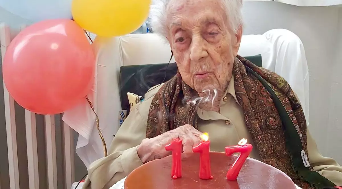 Maria Branyas Morera, pe scaun, cu un tort cu lumânări aprinse de culoare roșie care formează numărul 117, cu baloane roșii și galbene pe fundal