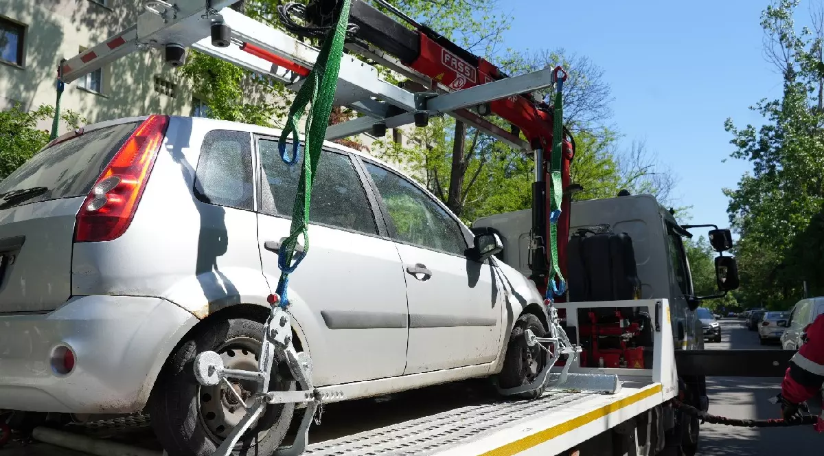 Imaginea arată o mașină argintie, un model de tip hatchback, care este ridicată și transportată pe platforma unei mașini de tractări. Mașina de tractări are un braț hidraulic roșu și verde, cu cabluri și sisteme de prindere specializate pe roți, folosite pentru a ridica și a asigura vehiculul. În fundal se pot vedea clădiri de locuințe și copaci, sugerând că acțiunea se desfășoară într-o zonă urbană, probabil o parcare sau o stradă. Vremea pare a fi însorită.
