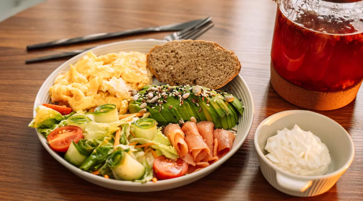 Pe o masă din lemn se află o farfurie cu un mic dejun. Meniul include ouă jumări, o felie de pâine neagră, salată verde cu roșii și castraveți, câteva felii de avocado presărate cu semințe, și bucăți de somon.Lângă farfurie se află o cană transparentă cu un lichid roșu, care ar putea fi ceai, și un bol mic, alb, cu o cremă, probabil brânză de vaci sau o altă pastă tartinabilă.