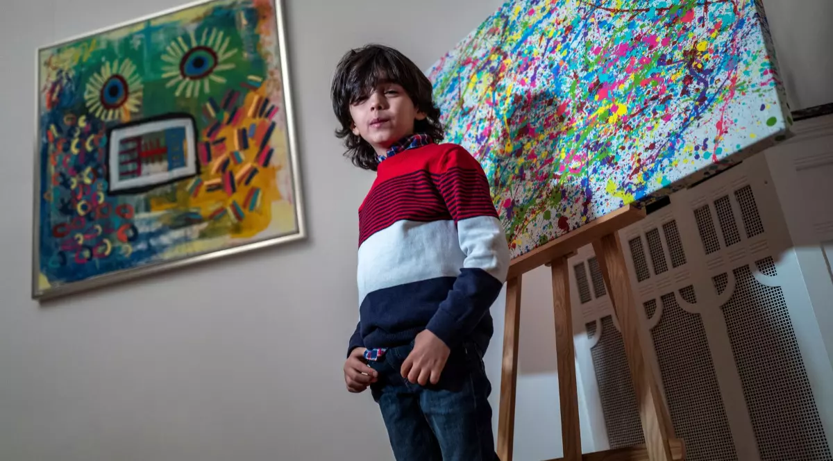 Mikail Akar îmbrăcat în blugi și bluză pozează în fața picturii sale „Champi” (dreapta) și a picturii sale „Sonnenblume Mensch” (stânga)