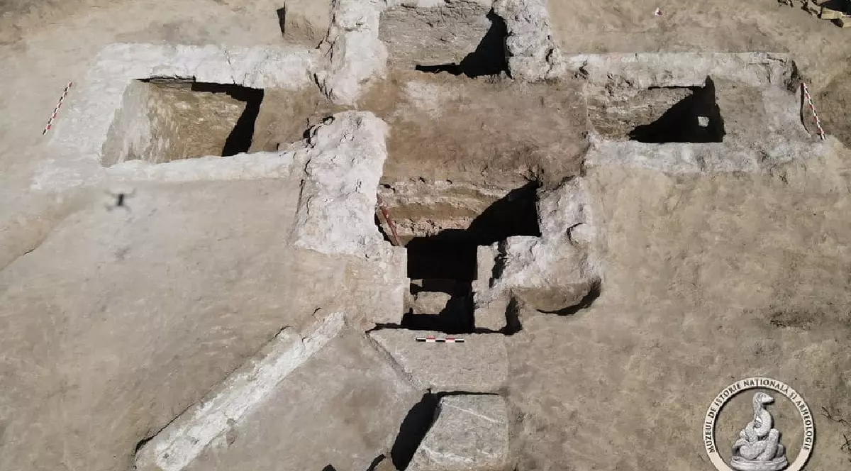 Imaginea, realizată dintr-o perspectivă de sus, arată un sit arheologic, probabil o descoperire recentă. Se observă o structură subterană din piatră, sub forma unei cruci sau a unei camere împărțite în mai multe compartimente. Pereții structurii sunt din piatră și pământ, cu o formă pătrățoasă. La baza imaginii, în colțul din dreapta, este vizibilă o siglă cu un șarpe, care aparține Muzeului de Istorie Națională și Arheologie Constanța.