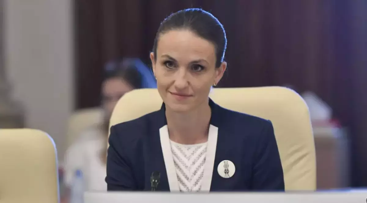 Oana Țoiu îmbrăcată într-un sacou negru și bluză albă în timp ce stă așezată pe un scaun