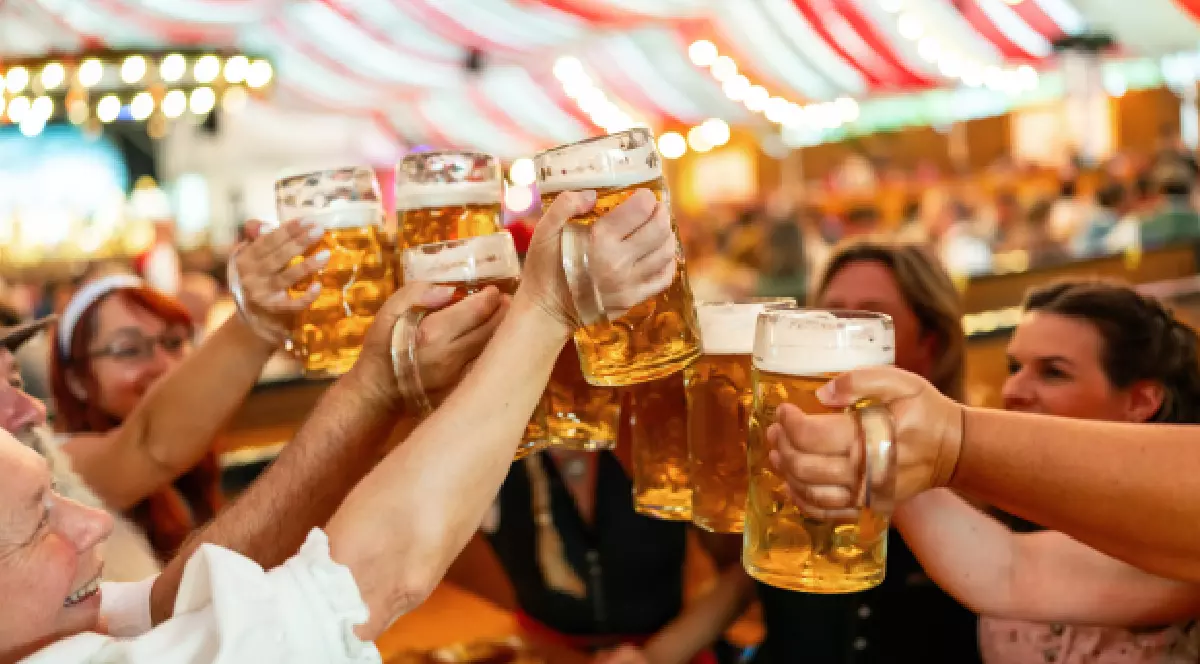 Imaginea surprinde un grup de oameni într-un cort de festival, ridicând halbe mari de bere pentru un toast. Atmosfera este festivă și plină de viață, cu tavanul cortului decorat cu dungi roșii și albe. Unii dintre participanți poartă haine tradiționale, cum ar fi o bluză albă, specifică festivalului Oktoberfest. Pe masă se poate observa un covrig mare, o gustare populară. În fundal se văd siluetele altor persoane, ceea ce sugerează că locul este aglomerat.