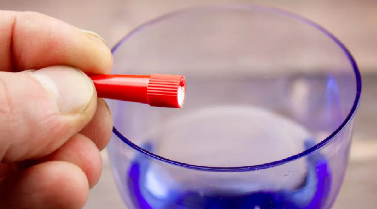 Imaginea prezintă o mână care ține un obiect mic și roșu, cilindric, ce pare a fi o capsulă sau un recipient. Obiectul este ținut deasupra unui pahar de culoare albastră, care conține lichid. Acțiunea sugerează o posibilă dizolvare a conținutului din obiectul roșu în lichid.