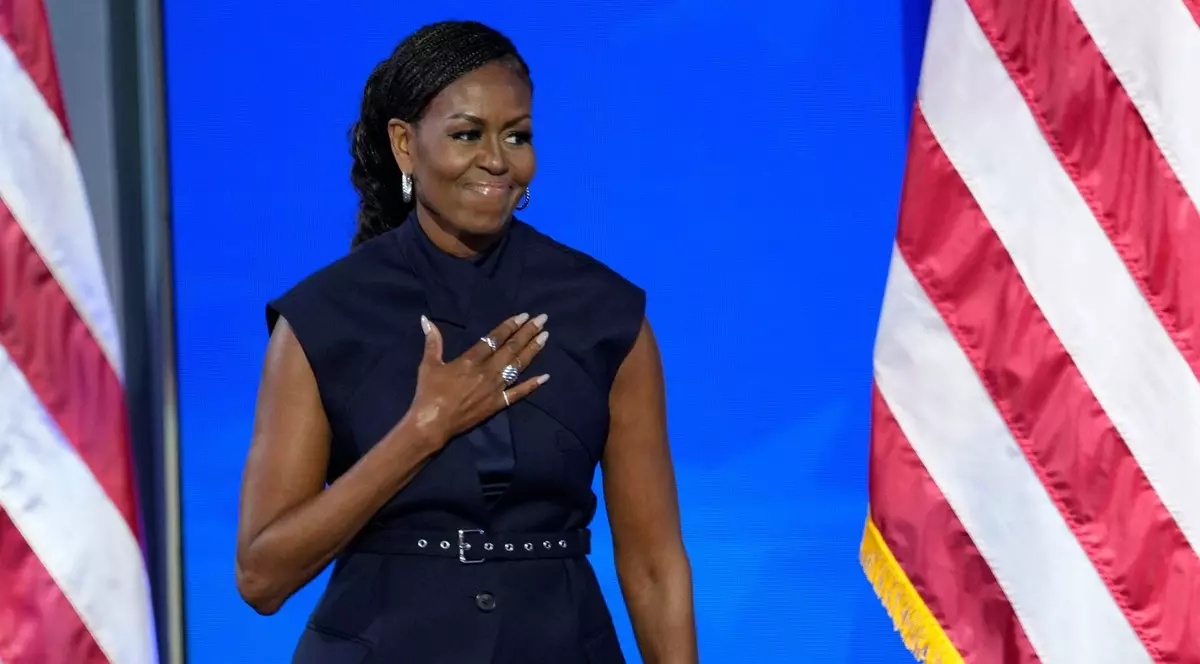 Fosta primă doamnă Michelle Obama, intr-o rochie neagra, fara maneci, vorbește pe scena ducand mana in dreptul inimii. in spate se poate observa steagul sua