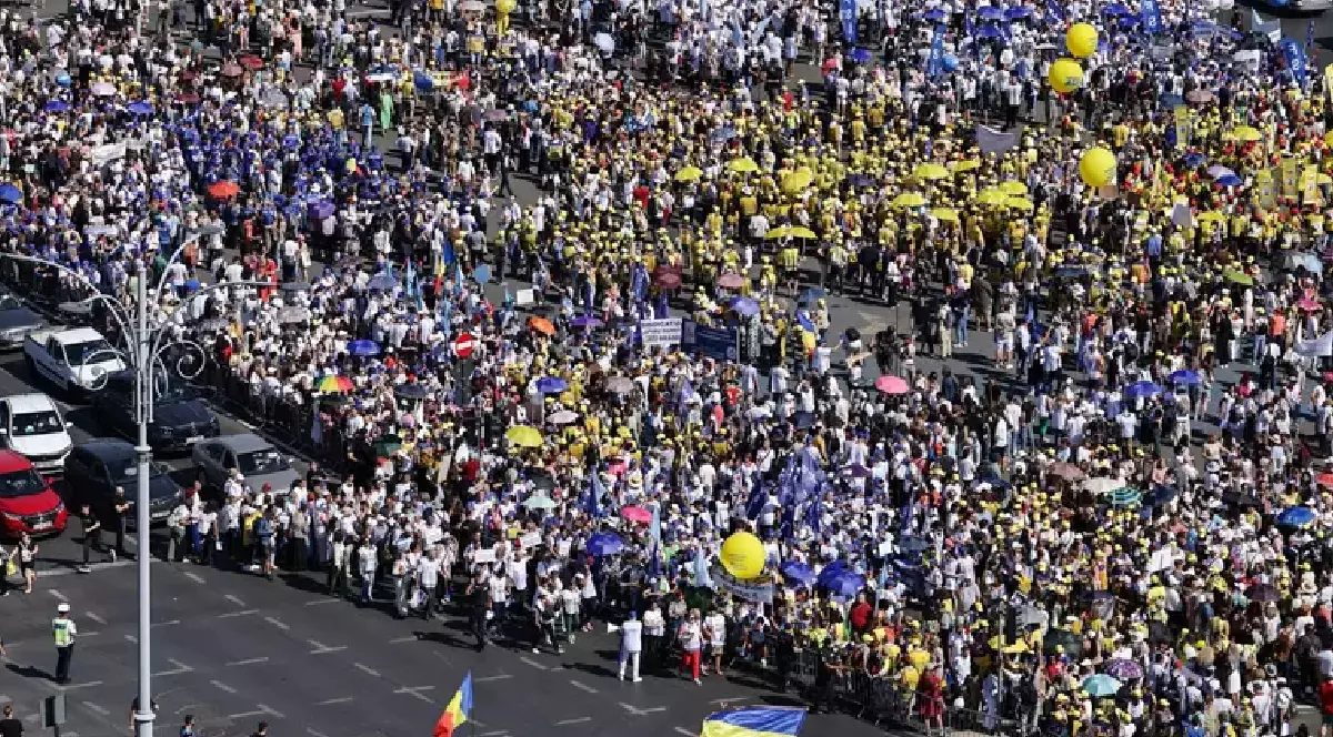 Fotografia, realizată dintr-un unghi înalt, protestul profesorilor care a avut loc în București. Mulțimea, formată din mii de oameni, poartă haine de diferite culori, dar predomină galbenul și albastrul, iar unii țin în mâini baloane galbene și umbrele.