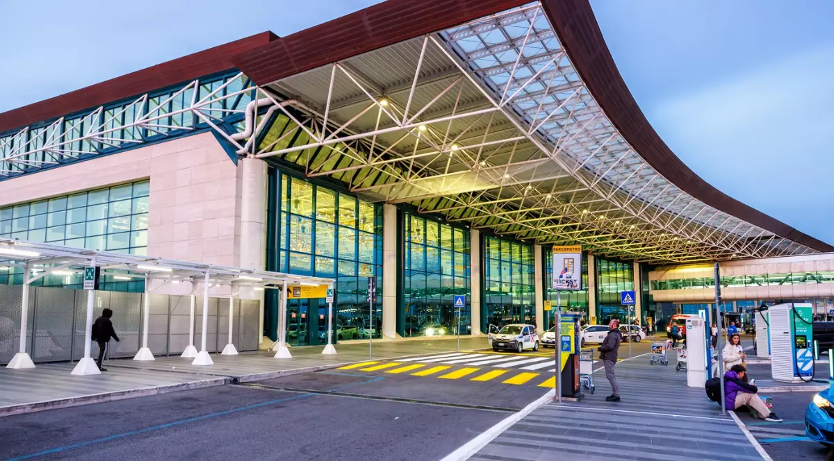 Imaginea surprinde intrarea modernă a unui aeroport la apus, cu o fațadă din sticlă și metal. Sub acoperișul curbat, câteva persoane așteaptă cu bagaje și cărucioare, în timp ce mașinile se deplasează pe banda de acces.