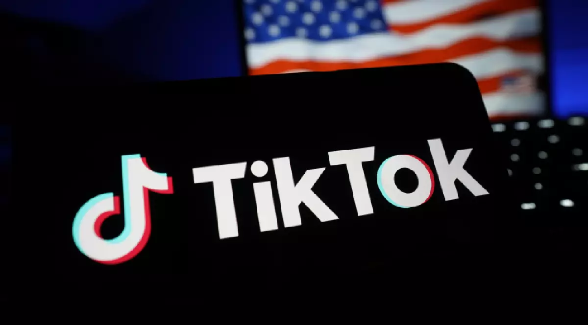 Imaginea surprinde logo-ul aplicației de social media TikTok afișat pe ecranul unui dispozitiv mobil, contrastând puternic cu fundalul întunecat. În fundalul nefocalizat, se distinge parțial un steag al Statelor Unite ale Americii pe ecranul unui alt dispozitiv.