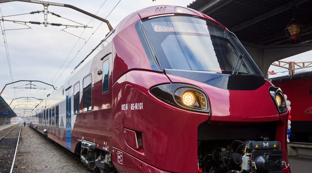 Fotografia surprinde un tren modern, de culoare roșie-grena, cu accente albe și gri, aflat într-o gară. Partea din față a locomotivei este rotunjită și are două faruri rotunde, cu lumina aprinsă. Pe laterale se pot observa vagoanele. Imaginea este realizată din unghiul șinelor, dând o senzație de viteză și dinamism.