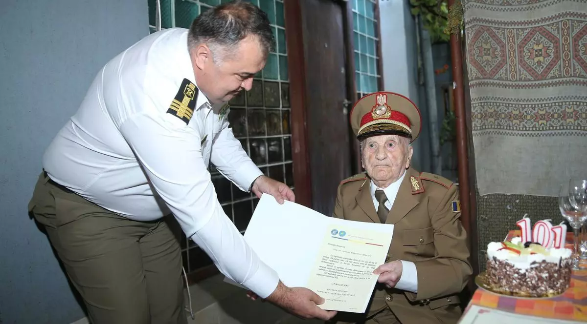 Veteranul de război Andrei Georgescu-Ionescu, la petrecerea de aniversare a vârstei de 101 ani, stând pe scaun îmbrăcat într-o uniformă militară lângă un tort cu candele cu cifra 101
