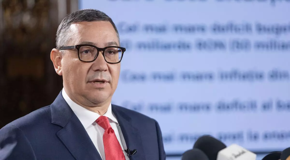 Victor Ponta, îmbrăcat în costum cu sacou de culoare albastru închis, cămașă albă și cravată roșie, iar pe fundal un panou pe care sunt cuvinte în blur