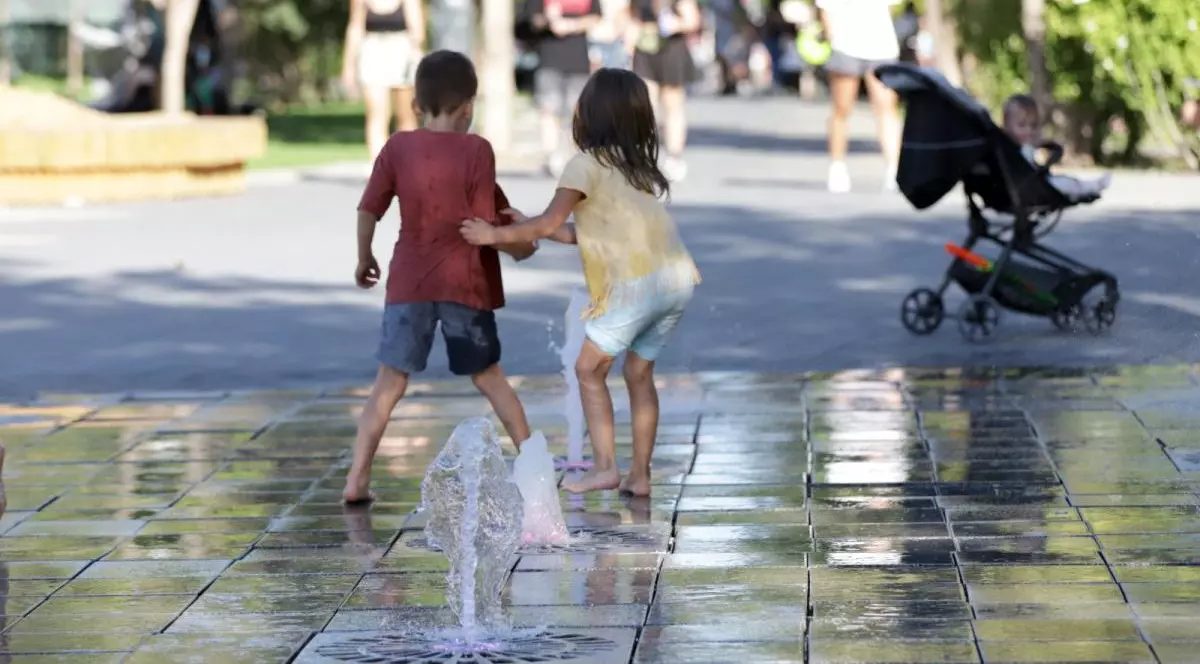 Doi copii, fotografiați cu spatele, se joacă în parc la o fântână de apă la sol, iar în jurul lor se văd oameni și un copil mic în căruț, București, 16 septembrie 2025