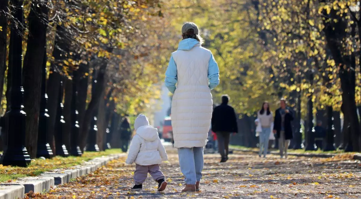 O mamă și un copil se plimbă în parc toamnă și sunt fotografiați cu spatele. Cei doi au geci, sunt acoperiți pe cap, iar pe jos sunt mutle frunze îngălbenite căzute