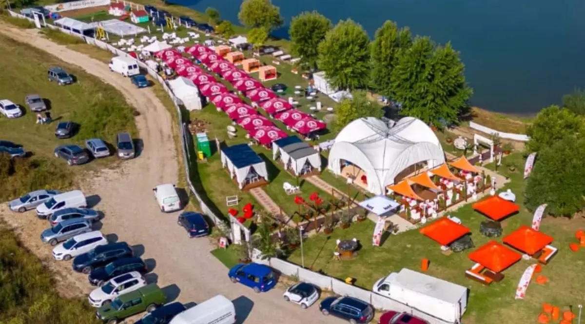 Imagine aeriană cu localul Arruba Garden din localitatea Paleu, Bihor, pe marginea unui lac, cu corturi și umbrele, și mai multe mașini parcate într-o parcare și pe un drum de acces