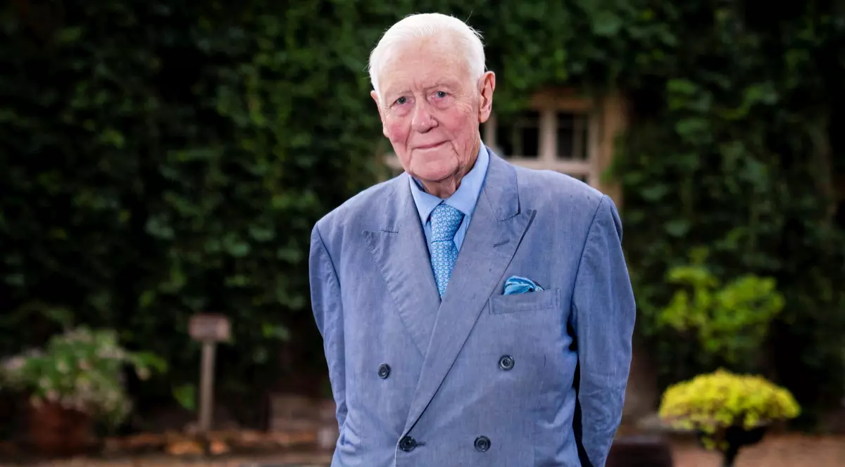 Benjamin Slade îmbrăcat în costum albastru deschis, în timp ce stă cu mâinile la spate