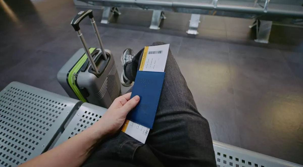 Calator sta asezat pe o banca in aeroport, picior peste picior, cu pasaportul si biletul de avion in mana stanga și un troller langa el