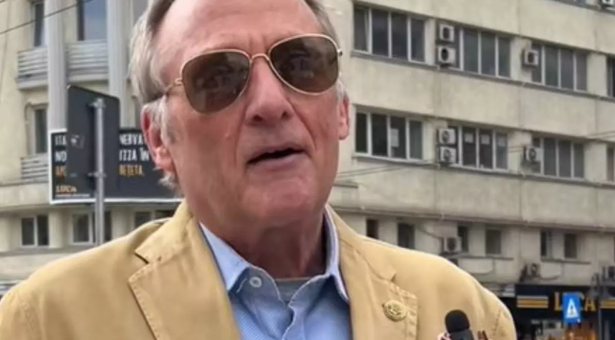 Un bărbat în vârstă, american, îmbrăcat cu un palton bej și o cămașa albastră, în timp ce acordă un interviu, n fundal se vede un bloc din centrul Bucureștiului