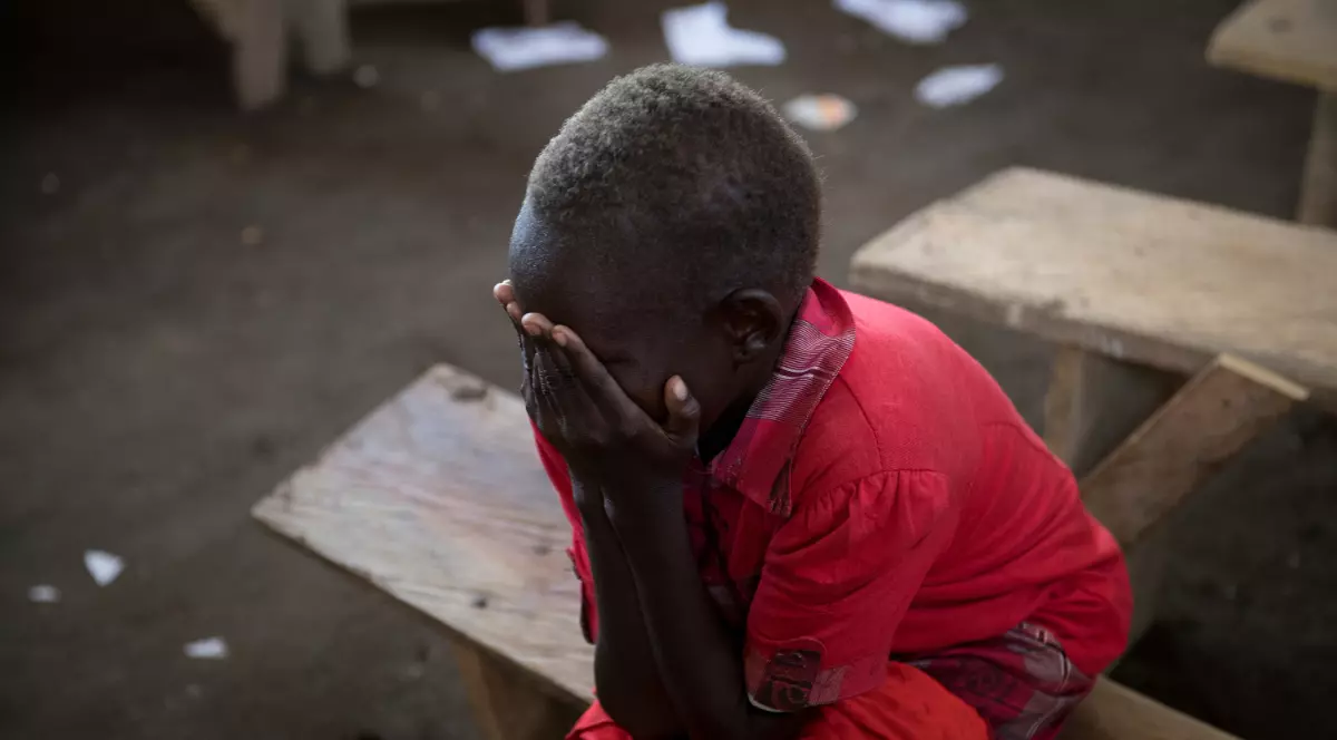 Imaginea surprinde un copil, îmbrăcat într-un tricou roșu, așezat pe o bancă simplă de lemn, cu fața acoperită de mâini, sugerând că plânge. Lumina slabă și fundalul întunecat și neclar, presărat cu hârtii aruncate, creează o atmosferă tristă și de disperare.