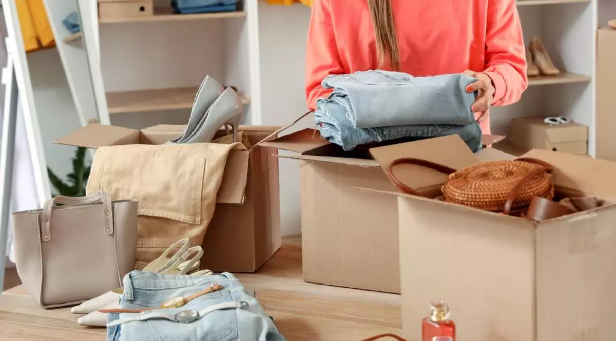 Femeie in bluza portocalie impacheteaza haine, genti si pentofi si le depozitează în cutii