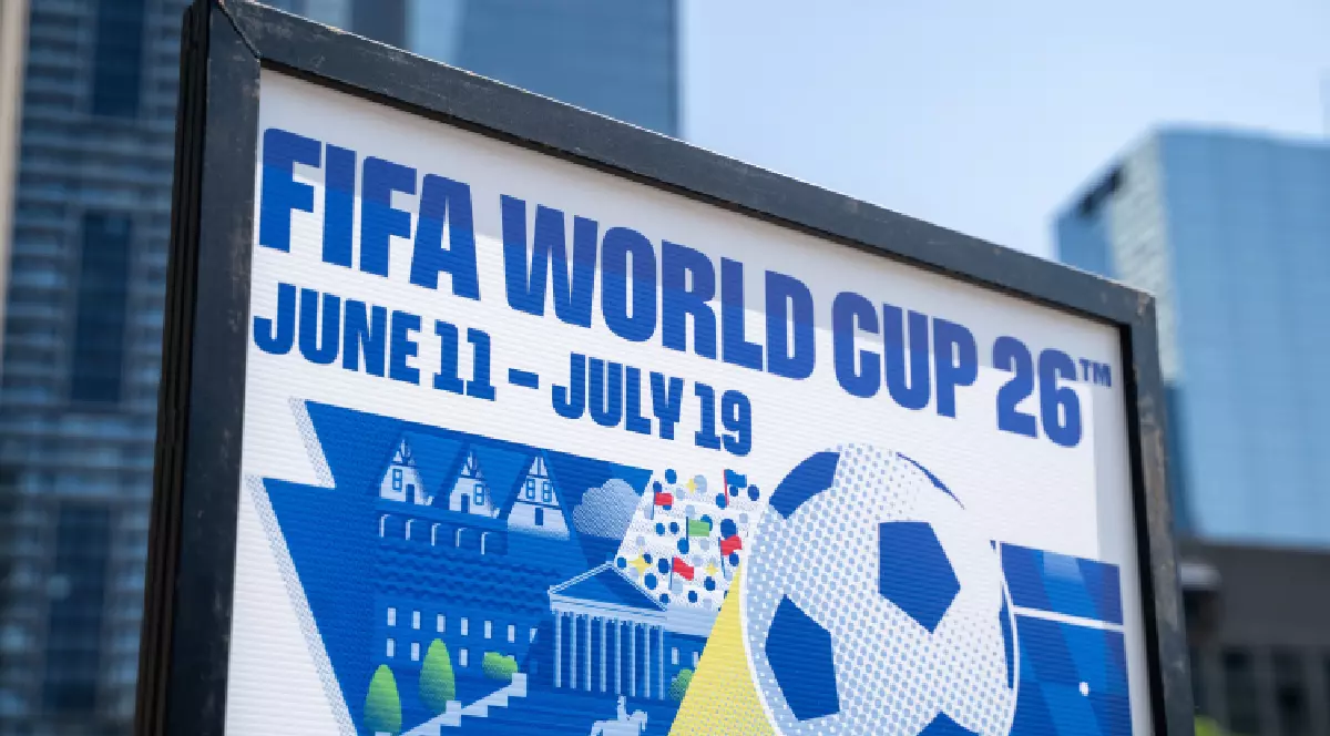 Imaginea prezintă de aproape un panou publicitar care anunță evenimentul sportiv FIFA World Cup '26 (Cupa Mondială 2026), specificând datele de desfășurare: 11 iunie - 19 iulie. Pe panou se află și o reprezentare grafică stilizată, incluzând o minge de fotbal și clădiri, iar în fundal se observă clădiri moderne de birouri.