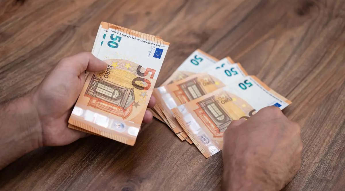 Imaginea surprinde un prim-plan al unei persoane care numără sau manevrează teancuri de bancnote de 50 de Euro de culoare portocalie, așezate pe o masă din lemn. Mâinile sunt vizibile ținând o parte din bancnote, în timp ce un alt teanc se află întins pe suprafața mesei.