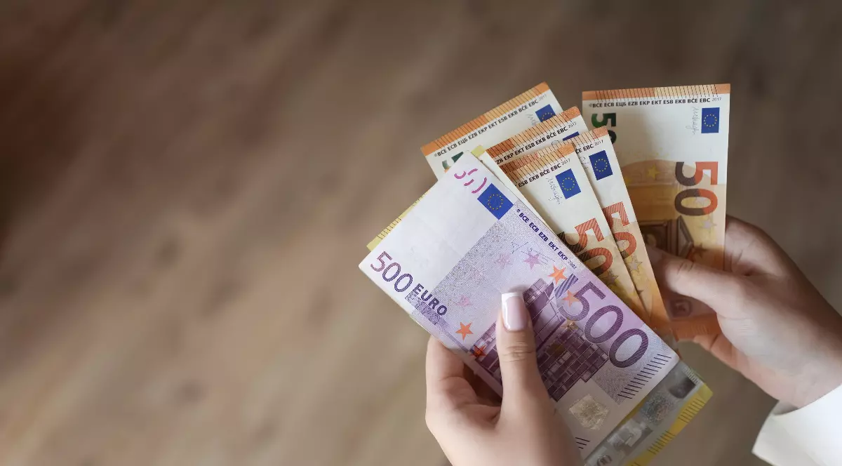 Imaginea surprinde un prim-plan al unor mâini care țin și numără mai multe bancnote Euro, inclusiv o bancnotă violet de 500 de Euro și bancnote portocalii de 50 de Euro. Mâinile au o manichiură îngrijită, iar fundalul este estompat și arată o suprafață de lemn.