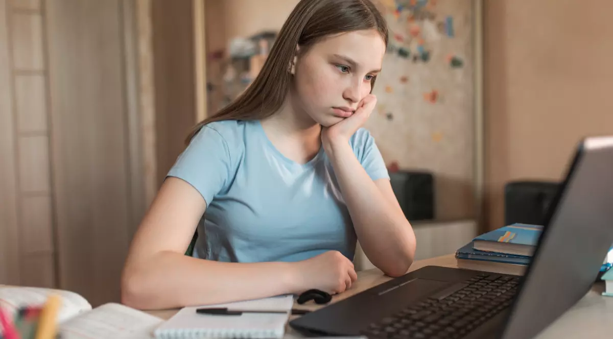 O tânără în tricou albastru deschis, care stă la un birou pe care se află un laptop și mai multe caiete