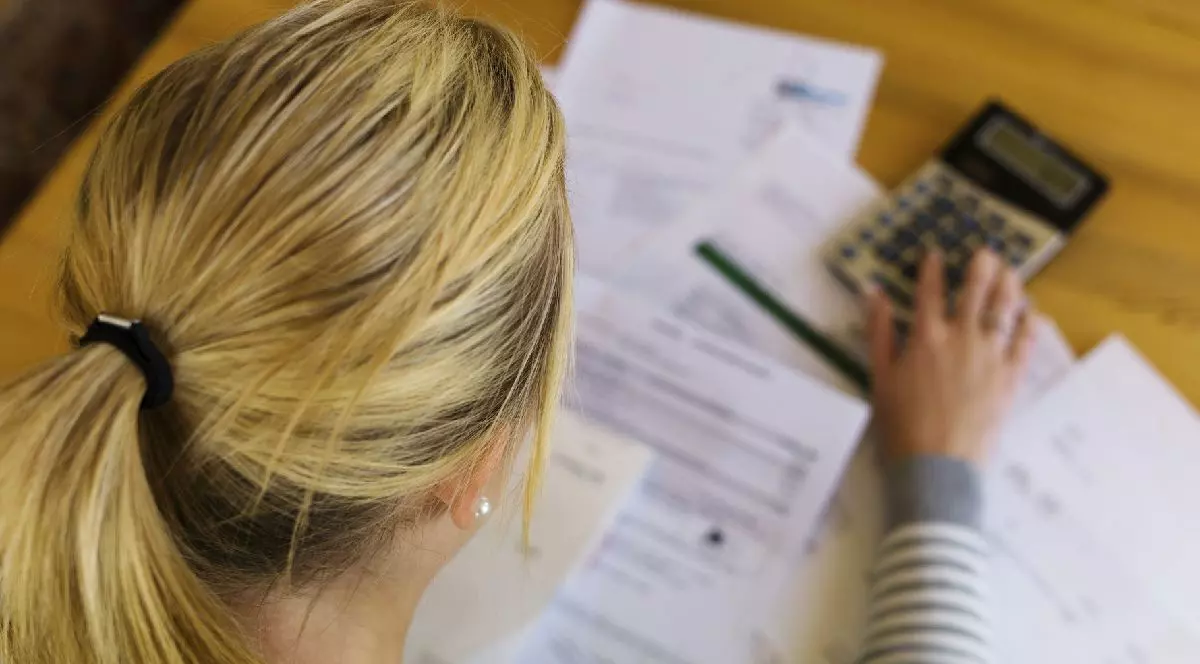 O femeie blondă, cu părul prins în coadă privește în jos, la facturile așezate pe birou. Imaginea ilustrează șomaj și faliment personal