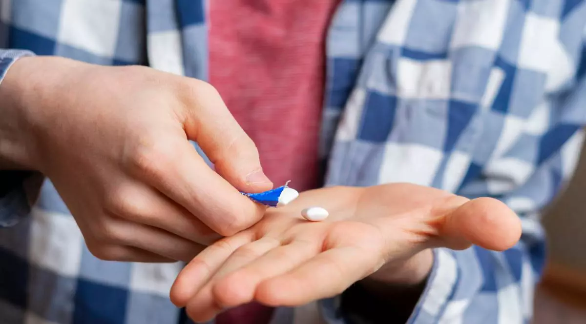 Un bărbat care are pune în palmă o gumă de mestecat dintr-un pachet, poartă o cămașă în carouri albastre