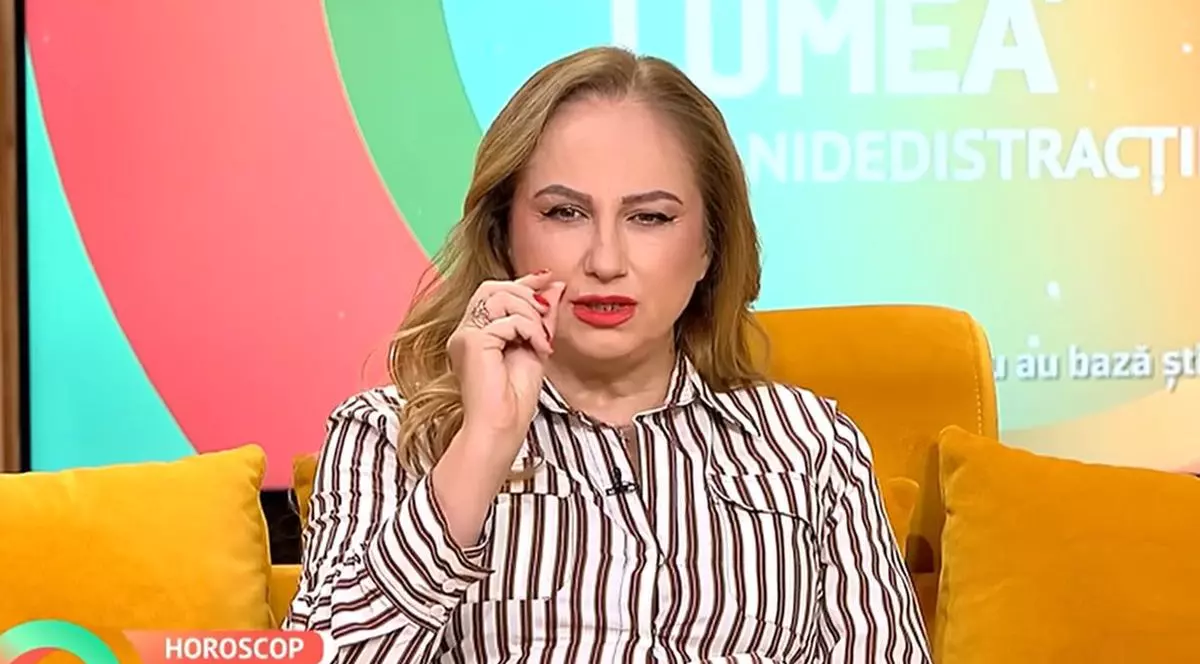 Astrologul Cristina Demetrescu, îmbrăcată cu o cămașă în dungi alb cu maro, vorbește și explică cu degetul despre ceva ce este mic, în timp ce stă pe o canapea portocalie în studioul unei emisiuni tv, luni, 6 octombrie 2025, București