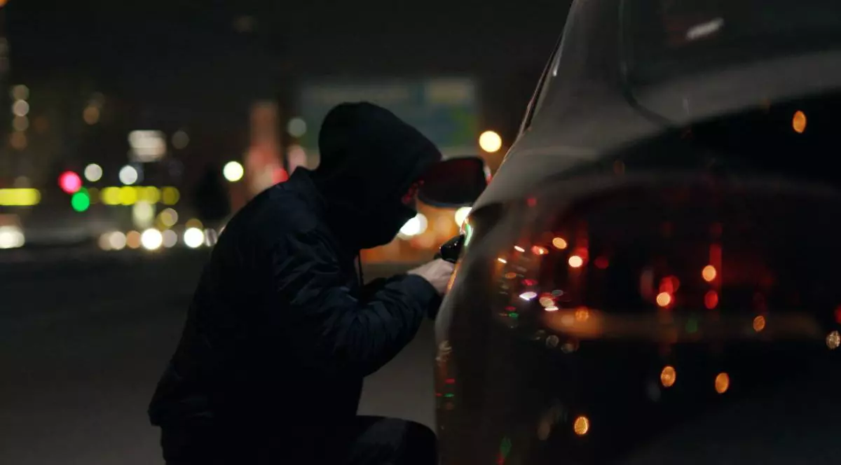 Un bărbat îmbrăcat în negru, cu gluga în cap, în timp ce încearcă să deschisă o mașină și să îi fure oglinda noaptea. Acesta folosește o șurubelniță