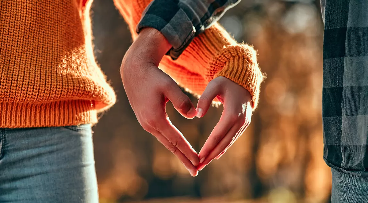 Imaginea surprinde mâinile unui cuplu care formează împreună simbolul unei inimi, evidențiind apropierea și afecțiunea dintre ei. Cei doi sunt îmbrăcați în pulovere sau cămăși de toamnă, iar lumina caldă a soarelui estompează frunzișul din fundal.