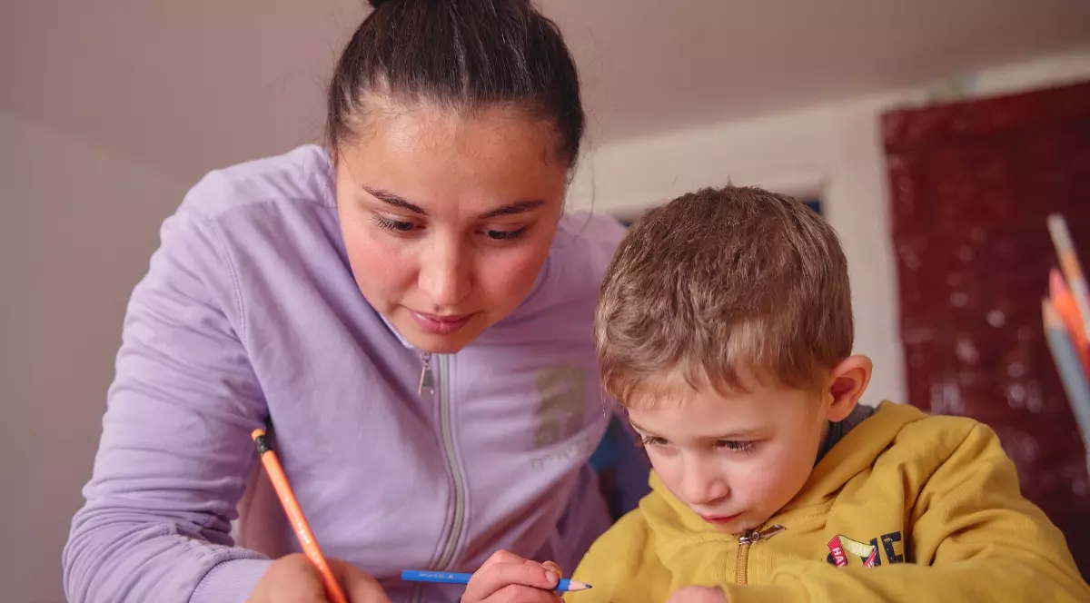 O mamă și fiul ei care sunt aplecați peste o masă și lucrează împreună la lecții. Amândoi folosesc creioane colorate, mama scriind pe hârtie în timp ce îl ghidează pe copil.