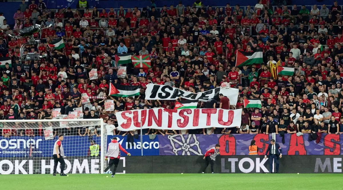 Imaginea surprinde un moment dintr-un meci de fotbal (posibil Osasuna-Getafe conform numelui fișierului), cu o tribună plină de suporteri îmbrăcați majoritar în roșu, care afișează diverse steaguri și bannere cu mesaje politice și locale. Printre steagurile palestiniene și basce, în centru se remarcă un banner mare cu textul "SUNTSITU" scris cu roșu pe fundal alb, sub un alt banner care pare să menționeze Israel.