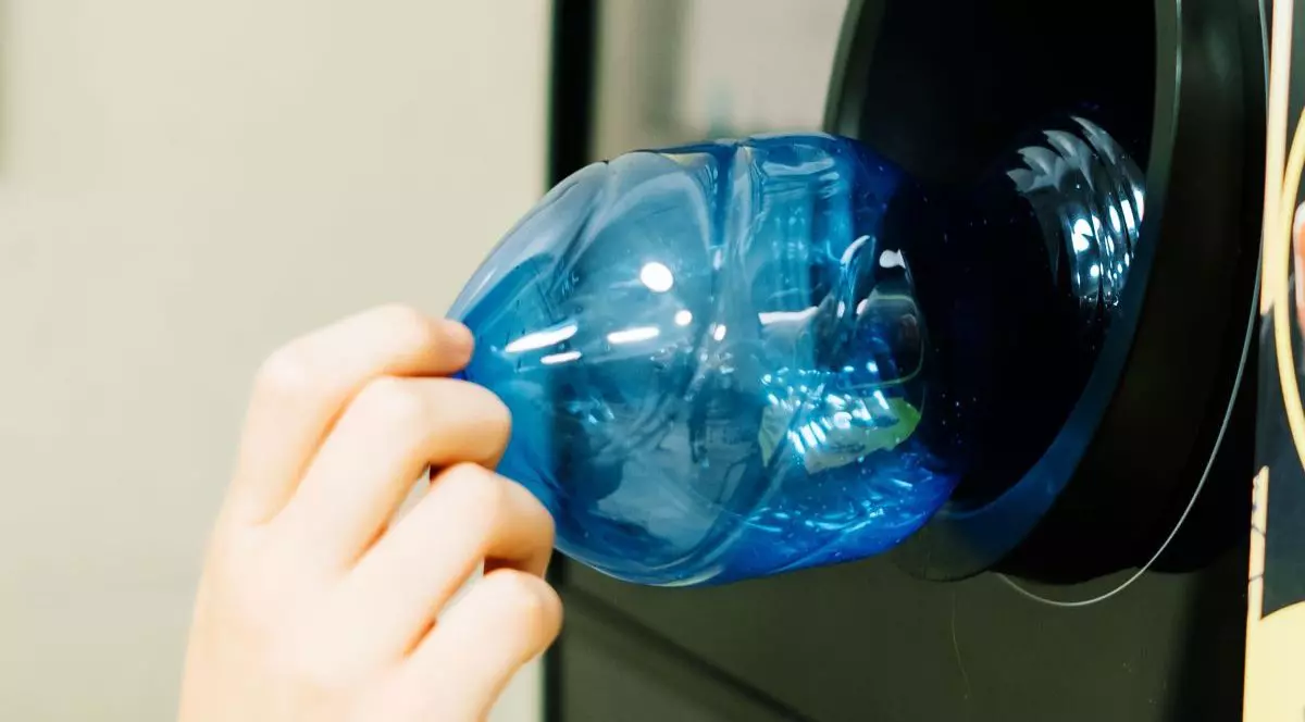 Mâna unui bărbat care bagă o sticlă albastră de plastic într-un automat de reciclare