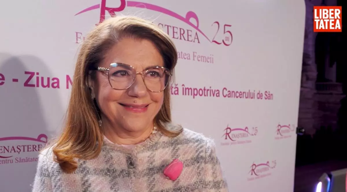 Mihaela Geoană, îmbrăcată într-o bluză roz cu dungi negre și o floare roz în piept, iar pe fundal un panou cu logo-ul Fundației Renașterea cu text roz pe alb