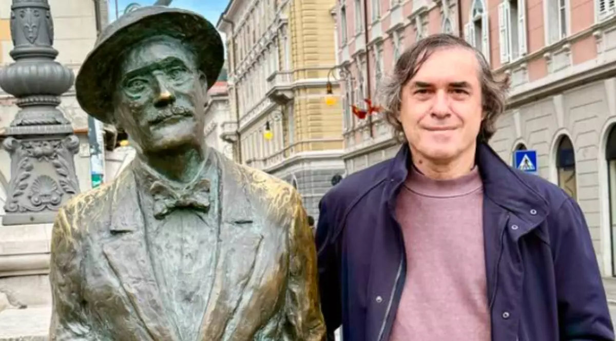 Mircea Cărtărescu îmbrăcat în bluză lila și geacă închisă la culoare, lângă statuia de bronz a lui James Joyce.